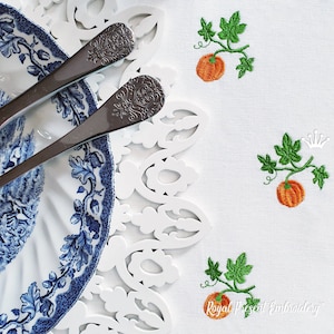 May include: White embroidered tablecloth with two orange pumpkins with green vines. The tablecloth has a white lace trim and a blue and white patterned plate with two silver forks on top.