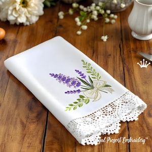 May include: White embroidered linen towel featuring a lavender and bow design. The embroidery includes purple lavender, green leaves, and a white bow. The bottom edge has a delicate white lace trim. The towel is folded on a wooden surface.