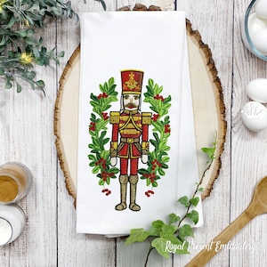 May include: Embroidered kitchen towel featuring a red and gold nutcracker soldier standing in a wreath of green holly and red berries. The text "Royal Present Embroidery" is at the bottom of the towel.