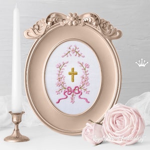 May include: A gold cross with a pink bow and floral wreath embroidered on white fabric in a gold oval frame. The frame is decorated with floral accents.