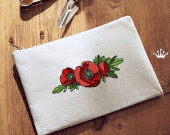May include: White canvas zippered pouch with a red poppy flower embroidery design. The pouch is on a wooden surface.