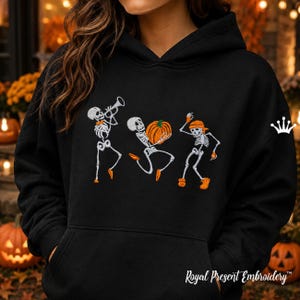 Könnte beinhalten: Ein schwarzer Kapuzenpullover mit einer Stickerei von drei weißen Skeletten. Ein Skelett spielt Trompete, eines hält einen orangefarbenen Kürbis und eines tanzt mit orangefarbenem Hut und Schuhen. Die Skelette haben orangefarbene Akzente.