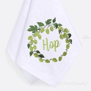 以下が含まれることがあります： 緑色のホップが刺繍された白いキッチンクロスで、中央に「Hop」という文字が刺繍されています。