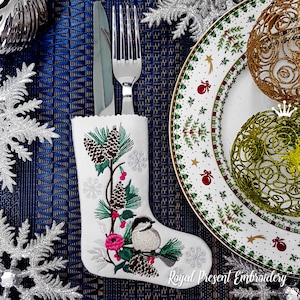 Puede incluir: Calcetín de Navidad blanco bordado con un pequeño pájaro, piñas y bayas de acebo. El calcetín está sobre un mantel individual azul con un copo de nieve plateado y un tenedor y un cuchillo.