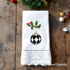 May include: White Christmas-themed hand towel with a decorative embroidered design. The design features a black and white diamond-patterned ornament, holly leaves, and red berries. The bottom edge has a white lace trim.