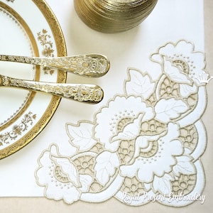 Könnte beinhalten: Ein weißer bestickter Tischläufer mit einem beigefarbenen Blumendesign. Das Design zeigt eine große Blume mit Blättern und eine kleinere Blume in der Ecke. Der Tischläufer liegt auf einer beigefarbenen Oberfläche.