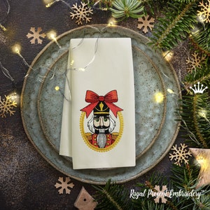 Könnte beinhalten: Ein weißes besticktes Küchentuch mit einer roten Schleife und einem Nussknacker-Motiv. Der Nussknacker trägt eine goldene und rote Uniform und hat einen großen Schnurrbart. Das Tuch liegt auf einem blauen und weißen Teller.