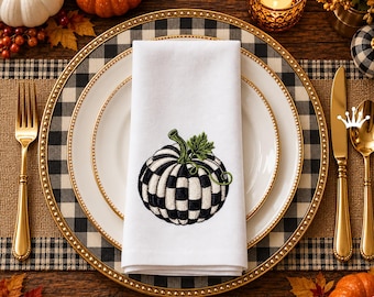 Puede incluir: Una servilleta de tela blanca con un diseño bordado de calabaza a cuadros blancos y negros con un tallo y hojas verdes, centrada en un plato llano blanco con borde dorado, sobre un plato de presentación a cuadros blancos y negros.
