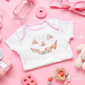 May include: A white baby onesie with pink polka dots and a floral patchwork pumpkin design. The onesie is on a pink background with other baby items, including a pink headband, a wooden toy, and a small white stuffed animal.