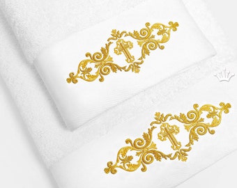 May include: Two white towels with gold embroidered cross designs. The designs feature ornate scrollwork and a cross in the center. The towels are folded and displayed on a white background.