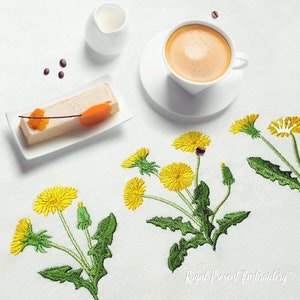 Pode incluir: Uma toalha de mesa branca com dentes-de-le&atilde;o amarelos bordados e folhas verdes. H&aacute; uma x&iacute;cara de caf&eacute;, um prato pequeno com uma fatia de bolo e um jarro de creme na mesa.