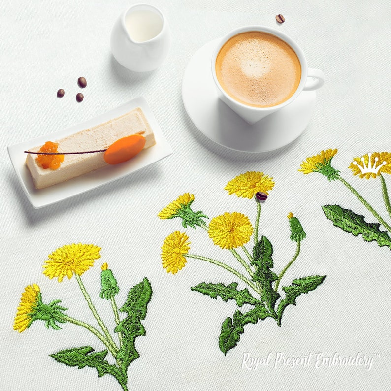Pode incluir: Uma toalha de mesa branca com dentes-de-le&atilde;o amarelos bordados e folhas verdes. H&aacute; uma x&iacute;cara de caf&eacute;, um prato pequeno com uma fatia de bolo e um jarro de creme na mesa.