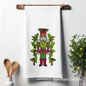 May include: White kitchen towel with an embroidered image of a Christmas nutcracker in a red and green uniform, surrounded by holly leaves and berries.