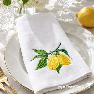 Könnte beinhalten: Weiße Leinen-Serviette mit gesticktem Design aus zwei gelben Zitronen und grünen Blättern. Die Serviette liegt gefaltet auf einem weißen Teller mit Goldrand. Der Text "Royal Present Embroidery" ist sichtbar.