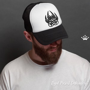 Peut inclure: Un chapeau de camionneur noir et blanc avec un motif d'empreinte de patte d'ours noir orné d'un nœud celtique. Le chapeau a un panneau avant blanc et un dos en maille noire.