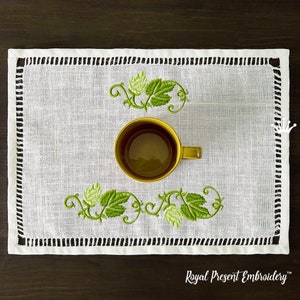 Pode incluir: Um sousplat de linho branco com um desenho de folhas bordadas em verde e uma borda decorativa. Uma caneca dourada está sobre o sousplat. O sousplat tem o texto "Royal Present Embroidery".