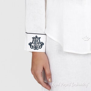 May include: White linen garment with a navy blue monogrammed cuff. The cuff features a decorative monogram in navy blue thread, framed by a thin navy border. The garment has a small crown emblem embroidered on the front.