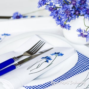 Könnte beinhalten: Eine weiße Serviette mit gestickten blauen Kornblumen, gefaltet und auf einen weißen Teller gelegt, darunter eine blau-weiß karierte Serviette. Auf der Serviette liegen eine Gabel und ein Messer. Im Hintergrund steht ein Strauß blauer Kornblumen.
