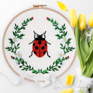 May include: Embroidered hoop art featuring a red ladybug with black spots surrounded by a wreath of green leaves and white daisies.