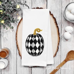 May include: A white kitchen towel with a black and white checkered pumpkin embroidered on it. The pumpkin has a gold stem.