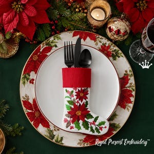 May include: A white ceramic plate featuring a red and green poinsettia pattern, topped with a white felt Christmas stocking cutlery holder. The stocking is embroidered with red poinsettia flowers, green leaves, and pinecones, and contains a black fork, knife, and spoon.
