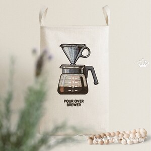 May include: A white canvas tote bag with a black and white illustration of a pour over coffee brewer. The text "POUR OVER BREWER" is printed below the illustration.