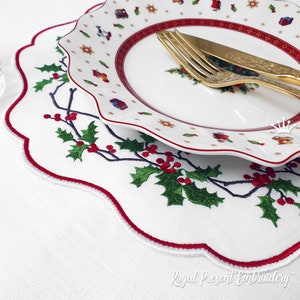 May include: A white embroidered placemat with a red border and a holly design. The placemat is set on a white tablecloth with a white plate and gold silverware on top.