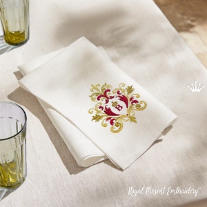 May include: Ivory linen napkins, folded and stacked, feature an embroidered crest with gold and burgundy accents. The crest includes a crown and the letter "B". The napkins are on a light beige tablecloth. The text "Royal Present Embroidery" is visible.