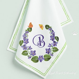 May include: White embroidered kitchen towel with a green border. The center features a wreath of purple flowers with a large purple letter "B" in the middle. Two small butterflies, one orange and one yellow, are above the wreath.