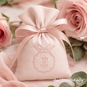 May include: A light pink drawstring bag with a bow closure. The bag features an embroidered design with the letter "R" inside a decorative frame. The background includes pink roses and the text "Royal Present Embroidery".