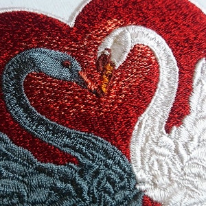Puede incluir: Diseño bordado de dos cisnes que se miran, formando una forma de corazón. El cisne de la izquierda es negro, y el cisne de la derecha es blanco. El corazón es rojo.
