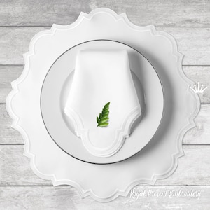 Può includere: Un tovaglietta bianca con bordo a festoni, un piatto bianco e un tovagliolo bianco piegato a forma triangolare. Una piccola foglia verde è posizionata sopra il tovagliolo. Il testo "Royal Present Embroidery" è visibile nella parte inferiore dell'immagine.