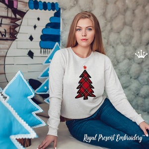 Op de afbeelding: Een wit sweatshirt met een rood en zwart geruit kerstboomontwerp. De boom heeft een rode ster bovenop. Het sweatshirt wordt gedragen door een persoon die op een vloer zit met blauwe kussens erachter. De tekst "Royal Present Embroidery" is zichtbaar onderaan de afbeelding.