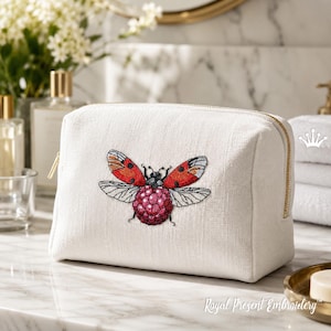 May include: A white cosmetic bag with a gold zipper and an embroidered ladybug design. The ladybug has red and black wings and a raspberry-colored body. The bag is made of a textured fabric.