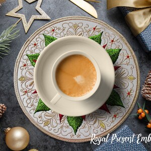 Peut inclure: Une tasse et une soucoupe &agrave; caf&eacute; cr&egrave;me reposent sur un dessous de plat d&eacute;coratif avec un motif floral et de feuilles. Le dessous de plat est entour&eacute; de d&eacute;corations de No&euml;l, de pommes de pin et d'un coffret cadeau. Le texte "Royal Present Embroidery" est visible.