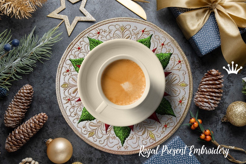 Peut inclure: Une tasse et une soucoupe &agrave; caf&eacute; cr&egrave;me reposent sur un dessous de plat d&eacute;coratif avec un motif floral et de feuilles. Le dessous de plat est entour&eacute; de d&eacute;corations de No&euml;l, de pommes de pin et d'un coffret cadeau. Le texte "Royal Present Embroidery" est visible.