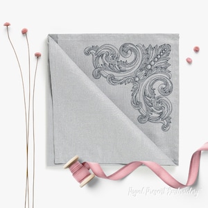 Pode incluir: Um guardanapo cinza prateado dobrado com um design de canto detalhado em cinza mais escuro. O design apresenta volutas ornamentadas e elementos florais. Uma bobina de fita rosa e flores secas também estão presentes.