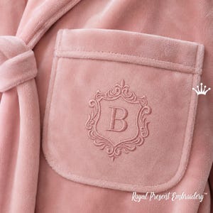 Könnte beinhalten: Nahaufnahme eines weichen, rosa Bademantels mit einer Tasche mit einem gestickten, verzierten Wappen mit dem Buchstaben "B". Der Bademantel hat einen passenden Gürtel und ist aus einem weichen, samtigen Material gefertigt. Der Text "Royal Present Embroidery" ist sichtbar.