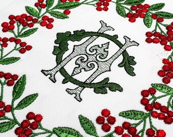 Könnte beinhalten: Gestickte Design mit einem Monogramm, umgeben von einem Kranz aus grünen Blättern und roten Beeren.