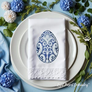 May include: A white napkin with a blue and white embroidered egg design. The napkin has a lace trim and is folded on a white plate.
