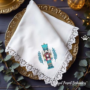 May include: White cloth napkin with a decorative embroidered nutcracker design. The napkin has a scalloped lace edge and is folded on a gold-colored plate. The nutcracker is in shades of blue, red, and tan.