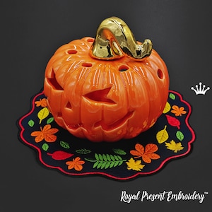 May include: A ceramic pumpkin with a gold stem, decorated with a carved jack-o'-lantern face, sits on a black fabric placemat with embroidered autumn leaves. The placemat has a red border.