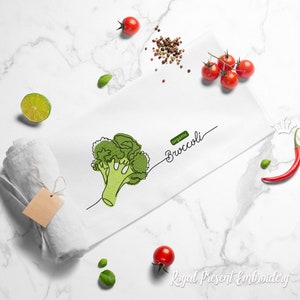 May include: A white kitchen towel with an embroidered image of a broccoli floret in green, and the text "organic Broccoli". The towel is rolled up and there are various vegetables and spices around it.