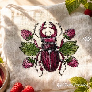 Könnte beinhalten: Besticktes Leinenkissen mit einem detaillierten Design eines Hirschkäfers, Himbeeren und grünen Blättern. Der Käfer ist weinrot, und die Himbeeren sind rosa. Der Text "Royal Present Embroidery" ist unten.