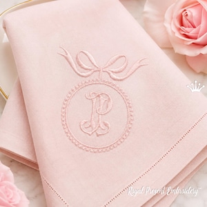 May include: Pale pink linen napkins with embroidered monogram letter "P" inside a circle, topped with a bow. The napkins have a delicate hemstitch border. The text "Royal Present Embroidery" is visible in the bottom right corner.