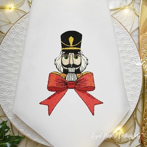 Puede incluir: Diseño bordado de un Cascanueces con un lazo rojo sobre un fondo blanco. El Cascanueces tiene un sombrero negro, una barba blanca y un bigote.