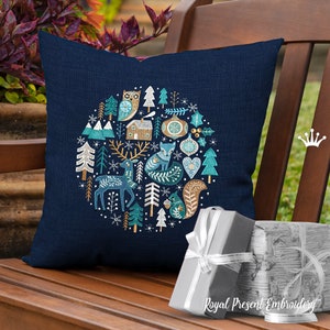 May include: A blue throw pillow with a circular embroidered design featuring a winter wonderland scene. The design includes a fox, deer, owl, squirrel, trees, and snowflakes. The pillow is on a wooden bench with a gift box in front of it.