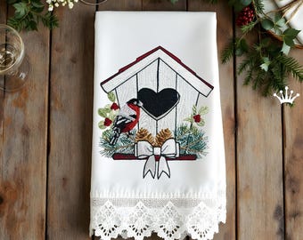 May include: White decorative towel with an embroidered birdhouse design. The birdhouse has a black heart-shaped opening, a red roof, and a bird. The design includes pinecones, greenery, and a white bow. The bottom edge has a white lace trim.