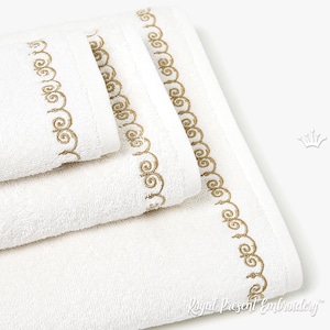 May include: A stack of three white towels with a gold embroidered border. The towels are folded and arranged in a pyramid shape. The embroidery is a delicate pattern of swirls.