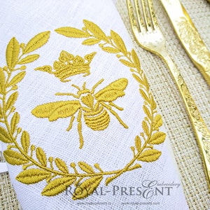 May include: A white linen napkin with a gold embroidered design of a bee and crown surrounded by a wreath of leaves. The text "ROYAL-PRESENT" is embroidered at the bottom of the napkin.